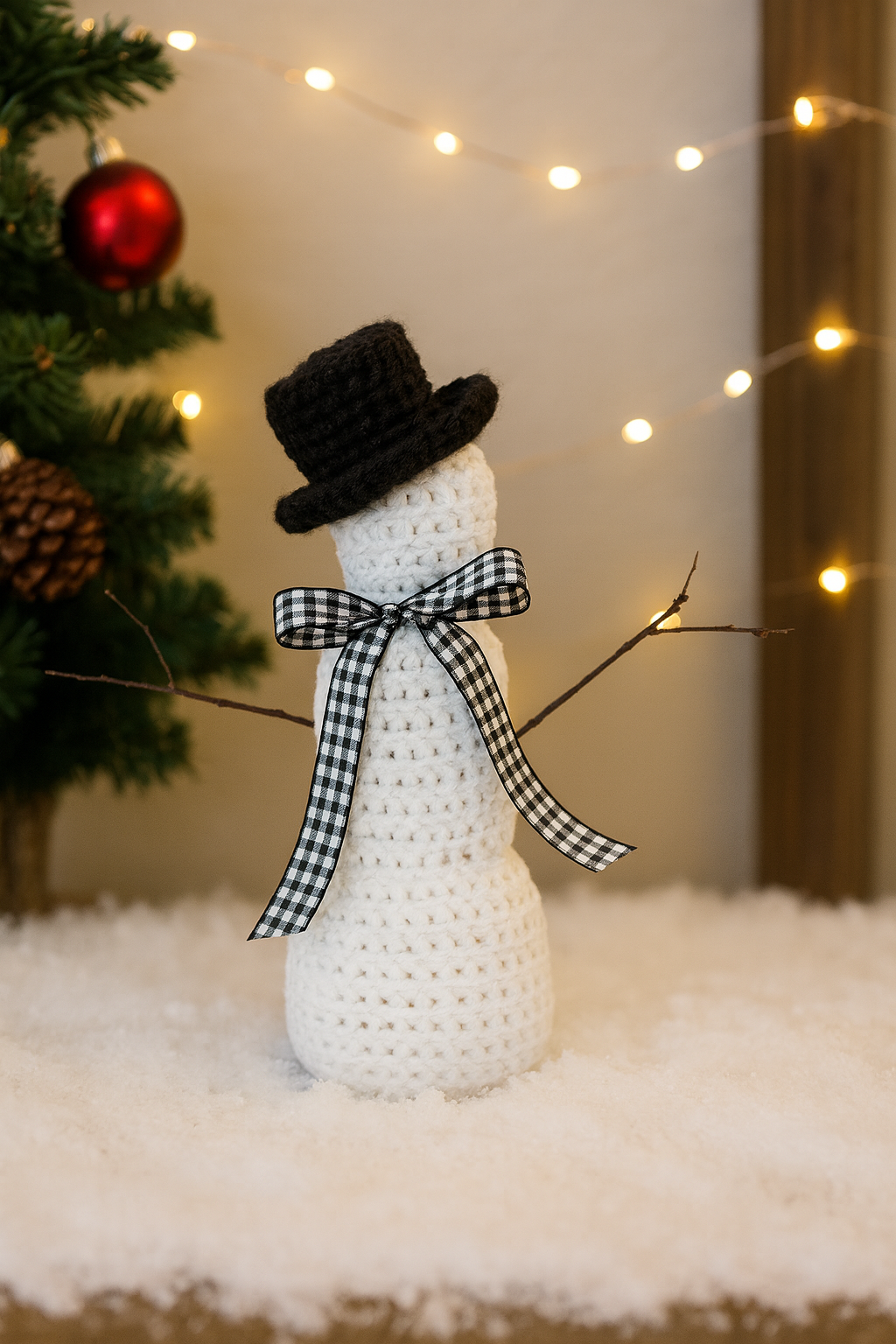 Crocheted snowman decoration with a black top hat and a checkered scarf in front of a Christmas tree.